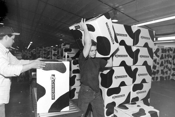 a black and white photo of two young men taking Gateway 2000 boxes off a conveyer and stacking them. The boxes feature the iconic black and white spotted cow print design of the time.