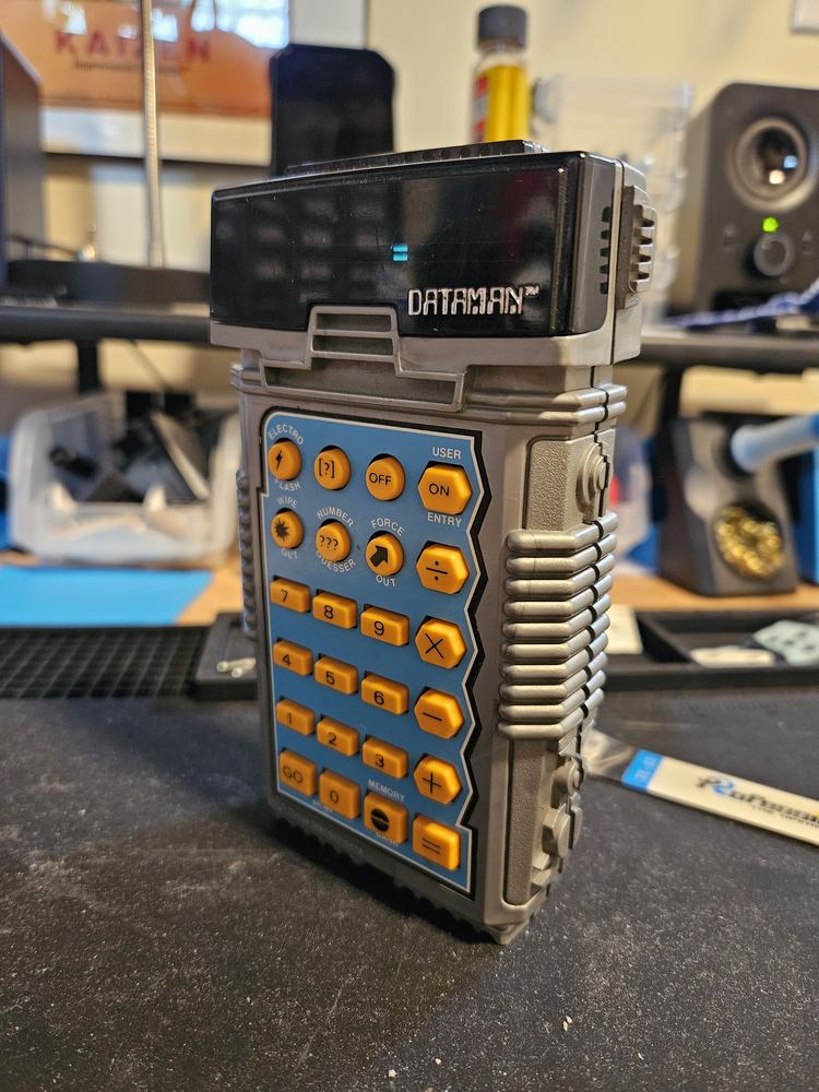 a toy calculator with a grey body and light orange buttons sits on tinsmith's workbench being tested. Pardon the case seam being half open, I forgot to tighten the screws before taking the picture.