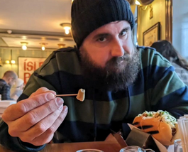 A bearded man wearing a black knit cap and a blue and green striped hoodie offering a small piece of fermented shark on a toothpick to the camera. Photo taken in Reykjavik, Iceland where fermented shark is a heritage dish.